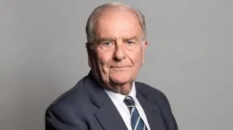 Sir Roger Gale looks at the camera. He has grey hair and is wearing a grey suit with a striped grey and lemon coloured tie. 