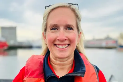 Picture of woman - Emma Wadee - smiling at camera, at sea, wearing bright clothing.