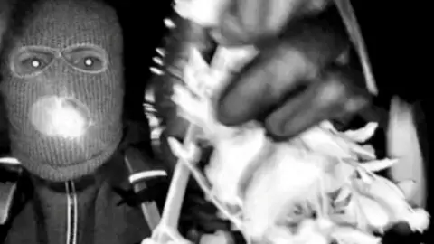Black and white security footage from Nancy Guthrie's home shows the suspect in the Guthrie kidnapping in a ski mask and a zipped up coat, wearing gloves and holding a flashlight in their mouth and holding branches being used to cover the lens. 