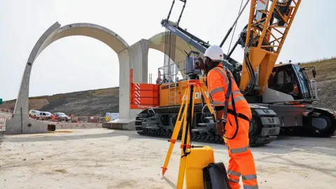 HS2 Ltd Two giant concrete arches on flattened ground with a surveyor in the foreground.