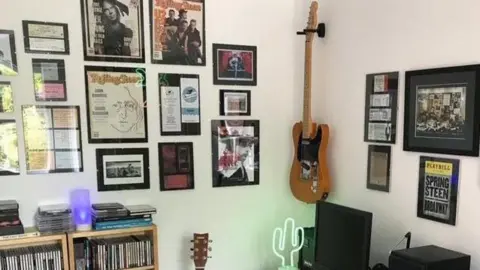 Phil Beard Frames with Rolling Stones magazine covers and other artwork can be seen on white walls. A rock guitar is pinned up in the corner and a shelfcase packed with singles stands beneath