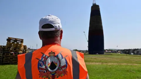 PA Media A man with an orange hi vis standing with his back to the camera in front of a large bonfire in Larne.