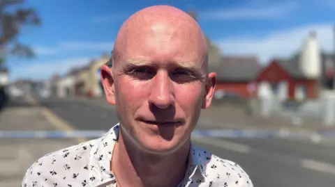 A man looks into the camera. He has a neutral expression. He is bald. He is wearing a white shirt with a blue pattern. In the background, which is blurred, we can see buildings lining a street. There is police tape across the road. It is a sunny day.