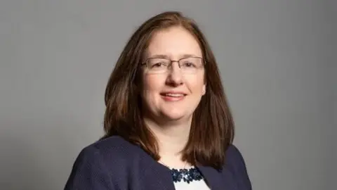House of Commons A head and shoulders view of a woman with brown, shoulder-length hair, taken in front of a grey backdrop. She is wearing rectangular glasses, a blue cardigan and a white top with floral design.