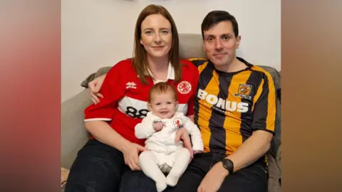 Supplied Emma and Simon Willingham are sitting side by side on a grey sofa with baby Beatrice smiling on their laps. Emma is wearing a red Middlesbrough shirt, and Simon is wearing a black and yellow Hull City shirt. They are all smiling.