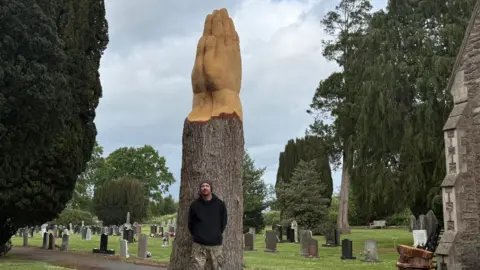 Tim Pearn Man in black clothes and hat stood in front of a tree truck. It has the prayer sculpture on top with two hands pressed together.