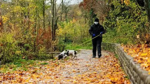 PA A man from Northamptonshire Police, dressed all in black holds a leash of a black and white police dog searching through a leafy path.