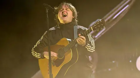 Jim Dyson/Getty Images Lewis Capaldi performing on stage with an acoustic guitar.