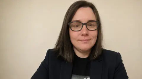 Peta Cooper Peta Cooper, with straight brown hair that sits on her shoulders, wearing black-rimmed glasses, and a black jacket over a black T-shirt