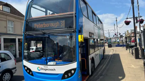 A Blue and white Stagecoach bus at a bus stop in the town of Ramsey,  a silver car is passing the bus and hanging baskets of colourful flowers can be seen on lampposts