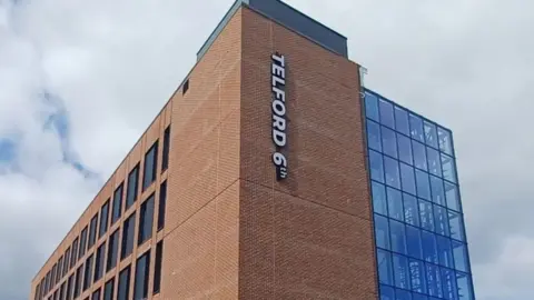 The top of a college building that has a tall red-brick tower and large glass window to the right of the tower. A sign reads "Telford 6th" vertically along the left of the tower. The other side of the building is also red brick with square black windows. The sky is cloudy in the background.