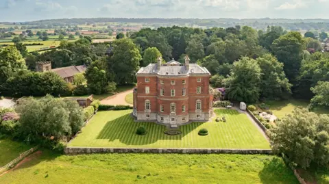 Mo Chaudry An aerial shot of a large hall with a large lawned area surrounding it. There is a church visible to the left hand side of the image and the buildings are set in rolling countryside with trees in the immediate background.