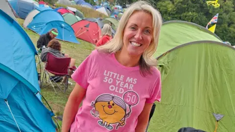 A woman with long blonde hair and wearing a pink t-shirt which says "Little Ms 50 year old" smiles as she stands in the middle of a campsite with tents behind her.
