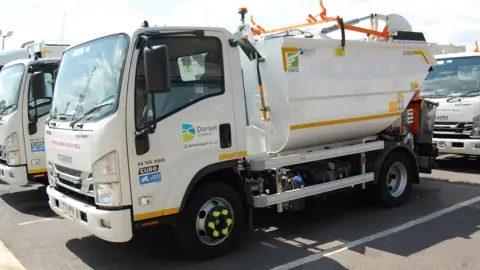 Dorset Council A small white food waste truck is parked near to others of its type in a car park. The Dorset Council logo is on the passenger-side door.