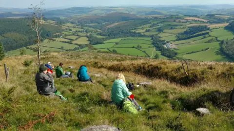 Kington Walks About five people sit on a grassy verge and look at the view of hills and trees in the distance.