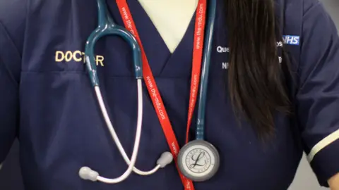 Getty Images A stock photograph of a doctor's chest. He is wearing a blue medical shirt. The word "doctor" is printed on it to the left and the NHS logo is on the opposite side. He is wearing a stethescope and some kind of lanyard around his neck. 