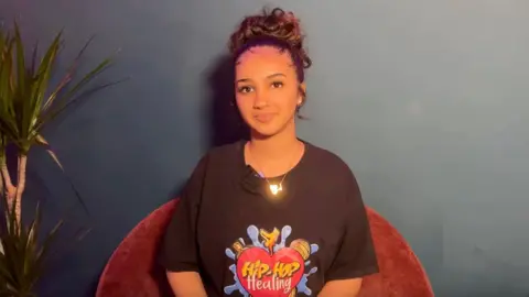 A picture of Rozaana sitting in a dark red chair in front of a dark blue wall, next to a plant on the left. She wears a shiny gold necklace and black shirt with a colourful Hip-Hop Healing graphic.