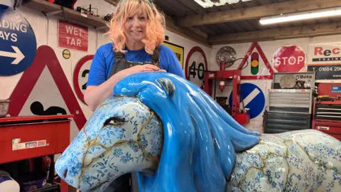 Jack Boskett Morris in black dungarees and a blue t shirt in front of the lion. Behind her is a studio with lots of different road signs in the background on the wall. 