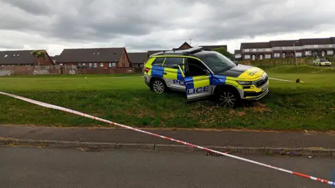 A wider shot of the abandoned police car on the grassy bank in a housing estate. Two long pieces of red and white police tape are cordoning off the vehicle. There is a another police car in the distance, parked closer to houses.