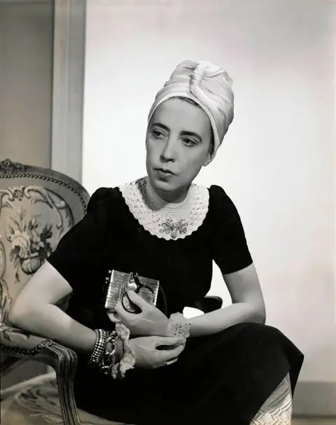 Getty Images A black and white image of Elsa Schiaparelli wearing a silk turban headpiece, a black dress with a white ruff around her neck. She is looking away from the camera and is also wearing a number of bracelets. 
