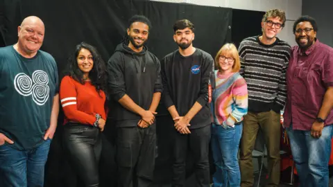 A group image of Emmerdale star Dominic Brunt, BBC New Comedy Awards Semi-finalist Seeta Wrightson, Coronation Street star Deborah McAndrew, Emmerdale star Mark Charnock, and director of the play Dermot Daly, with young actors from Bradford Solomon Gordon and Armaan Ali in the centre.