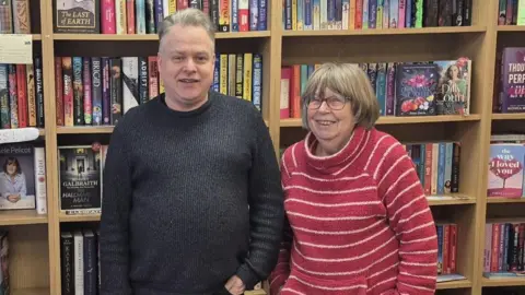 An older man and woman stand side-by-side in front of a bookshelf filled with colourful books. The man has short grey hair 