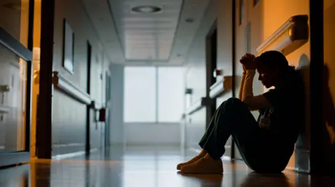 A female staff worker sits on the floor of a hospital corridor with her head in her hands. She is in the shadows