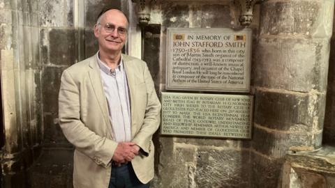 Adrian Partington, who is wearing jeans and a cream blazer, standing in front of a plaque to John Stafford Smith inside Gloucester Cathedral
