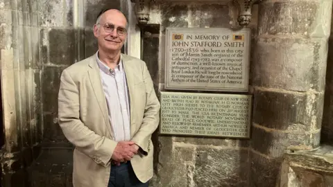 BBC 的阿德里安·帕丁顿身穿牛仔裤和奶油色西装外套，站在格洛斯特大教堂内纪念约翰·斯塔福德·史密斯的牌匾前
