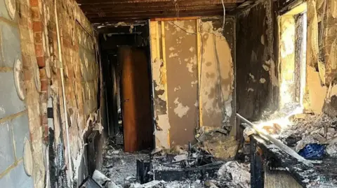 Suffolk Fire and Rescue Service The picture is taken of a room that has got severe fire damage. The walls are stripped, and are now brick. On the right there is lots of black burnt pieces of wood and materials scattered. 