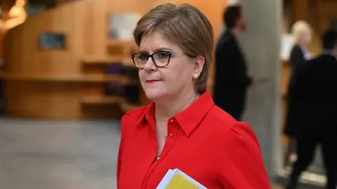 Nicola Sturgeon, wearing a red shirt and glasses, carries a yellow folder under her left arm while walking indoors 