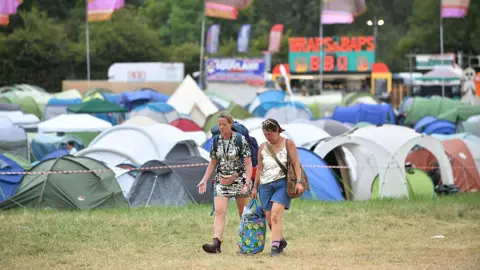 Getty Images Two women are walking alongside tents in a field, carrying bags. Food stalls and festival flags can be seen in the background.