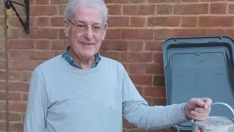 A man standing in front of a brick wall. He is wearing a pale blue jumper and checked shirt. He has grey hair and is also wearing glasses. He has a tin of paint in his left hand.