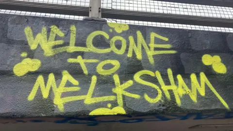 A wall with yellow graffiti on it reading "Welcome to Melksham".