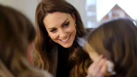 The princess seated at a table with two children, who are positioned in the foreground with their backs and profiles visible. Catherine has long, wavy hair that falls over their shoulders and appears to be engaged in a calm, focused interaction with the children. One of the children is holding a seashell to her ear.
