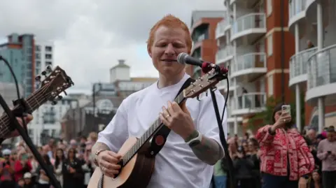 Ed Sheeran wearing a white t-shirt playing guitar and singing in to a microphone on the Ipswich waterfront, crowds of people can be seen in the background