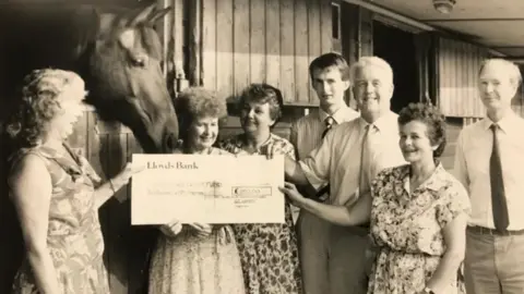 Gibbs family A group holding a cheque in stables by a horse