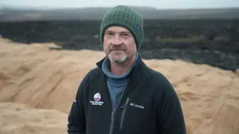 BBC/Stuart Prandle A man, Miles, looks into the camera. In the background, a moorland is blackened apart from a light brown mat covering some of it.