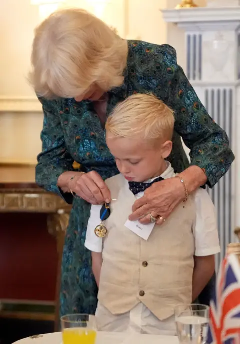 PA Media Rupert Williams standing in front of Queen Camilla. Rupert wears a white short sleeved shirt, a beige waistcoat and a black bow tie. Queen Camilla wears a patterned green, blue and orange dress. The Queen is pinning the medal on him