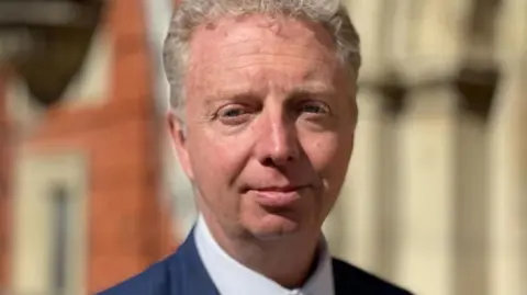 Alex Allinson, who has short wavy light coloured hair and is wearing a blue suit jacket over a shite shirt. He is standing in front of the House of Keys public entrance.
