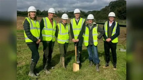 Six people in yellow high vis jackets and white hard hats on a field. One of the people in the middle is holding a shovel.