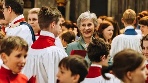 Gloucester Cathedral The Duchess of Gloucester's head and shoulders can just be seen in the middle of the picture surrounded by lots of choristers of all ages inside the cathedral. She is smiling to a young man to the left, who is wearing red and white robes. 