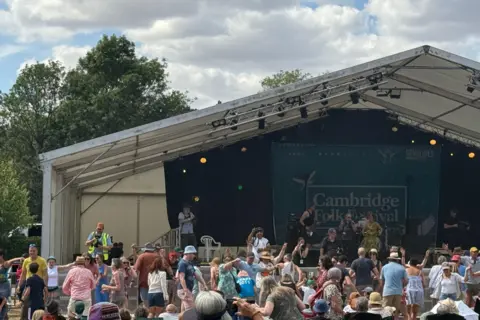 Mark Walsh/BBC Crowds sit and stand in front of a stage. A banner is unfurled at the back of the stage, reading CAMBRIDGE FOLK FESTIVAL. It is cloudy above with some patches of blue sky. 