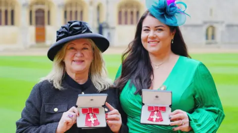 Alison Madgin on the left is smiling and wearing a black coat and stylish hat. Carly Barrett is wearing green, long-sleeved dress with low neckline and a blue fascinator-style hat. They are holding up their boxed honours in the shape of a red bow. 