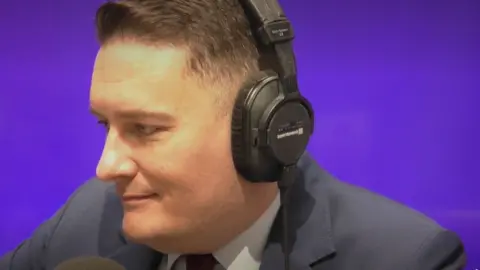 A man, Wes Streeting, wearing a suit and a set of over ear headphones. He is sat in a room with a purple background.