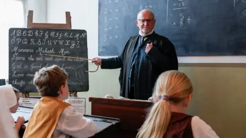 National Lottery Heritage Fund A man dressed in teachers robes points at blackboard with a cane.
The blackboard has old fashioned style writing in chalk on it.
Two children sit at wooden desks looking at him.