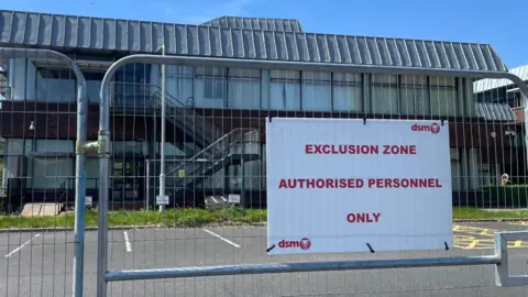 A two-storey building with large glass windows is seen behind a security fence which has a sign on it that reads "exclusion zone" and "authorised personnel only" in red letters on a white background. There are car parking spaces alongside the building.