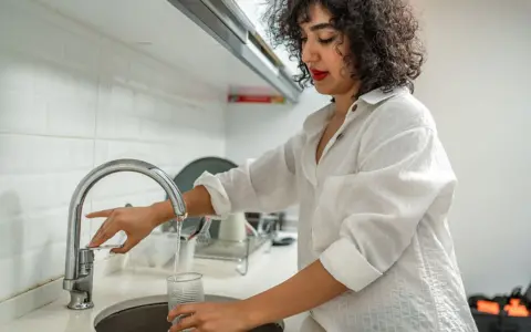 Getty Images A woman fills a glass with water from a kitchen tap at home, she's wearing a white shirt and the kitchen has white tiles and a dish drying rack just visible behind the sink.