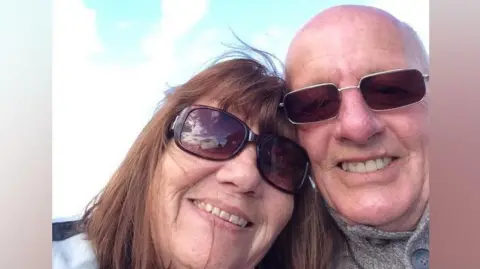 Kathryn Stephenson and Chris Belshaw are taking a selfie and smiling at the camera. Both of them are wearing brown sunglasses. There is a cloudy blue sky behind them. She has shoulder-length brown hair with a fringe and he is bald with light cropped hair at the sides.