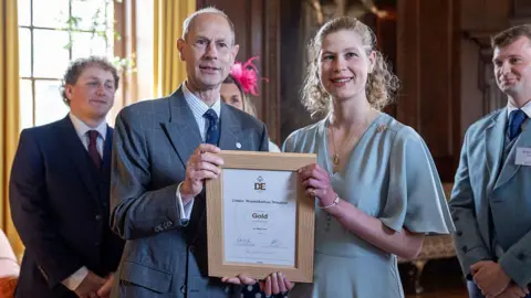 Getty Images Prince Edward, Duke of Edinburgh presents his daughter Lady Louise Mountbatten-Windsor with the Duke of Edinburgh's Award Gold Award at Palace of Holyroodhouse. They are dressed formally and standing side-by-side, smiling. The award is a document in a wooden frame.